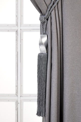 May include: A silver tassel tieback with a twisted metal design. The tassel is attached to a gray rope and is hanging from a gray curtain.